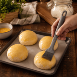 Grey silicone brush with ridged bristles being used to apply egg wash to buns.